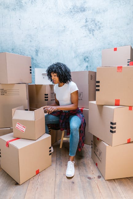 A young woman with curly dark hair, wearing a white t-shirt, blue jeans, and white sneakers, is sitting on a wooden chair amidst numerous large cardboard moving boxes in a room with a textured light b
