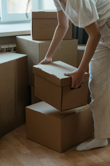 A young woman with curly dark hair, wearing a white t-shirt, blue jeans, and white sneakers, is sitting on a wooden chair amidst numerous large cardboard moving boxes in a room with a textured light b